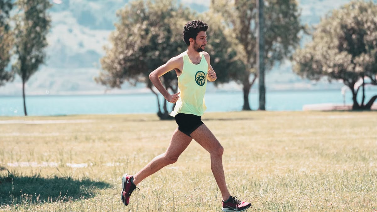 Side view of young determined male athlete in sportswear running on lawn in park near lake in sunny morning